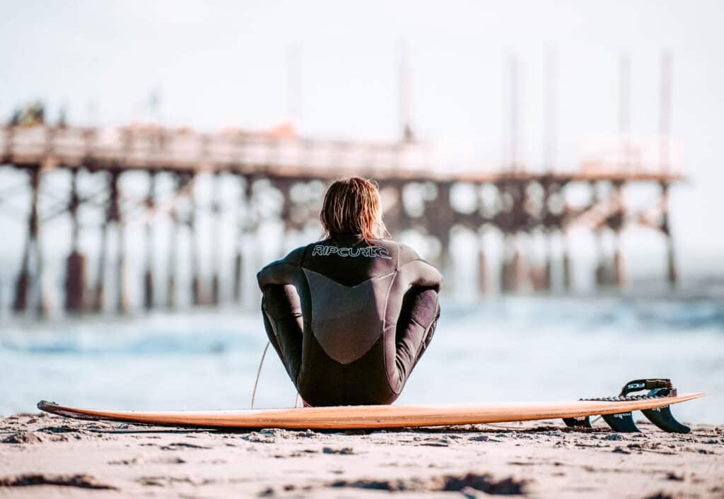 A person sitting on top of a surfboard at the beach looking at a pier in the distance