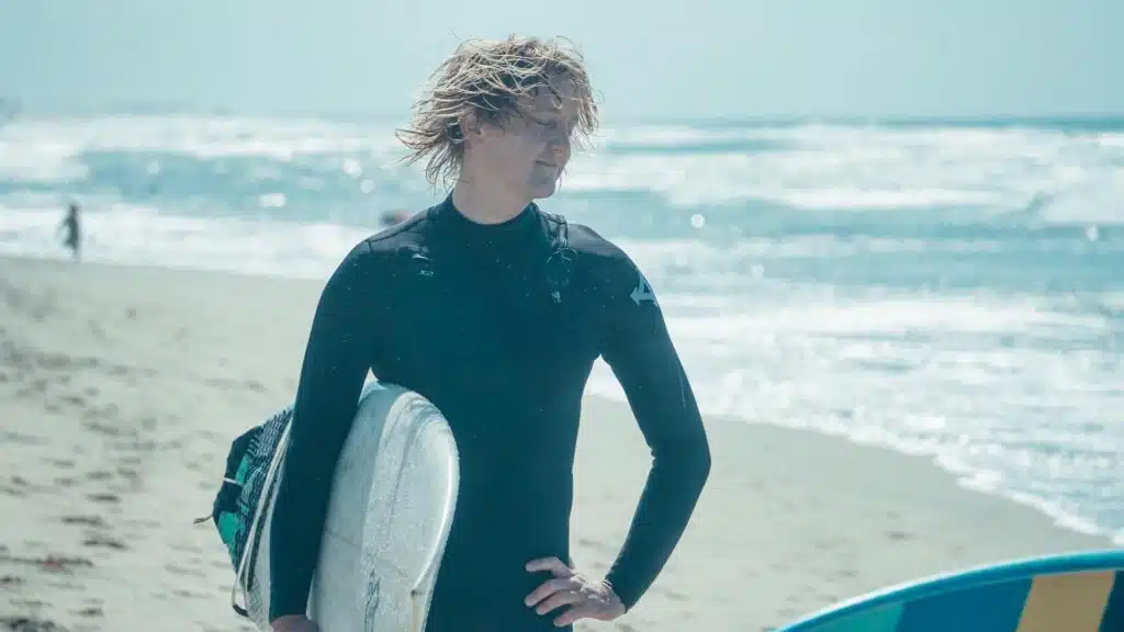 Ein Bild eines Mannes, der ein Surfbrett hält, er hat unordentliches blondes Haar