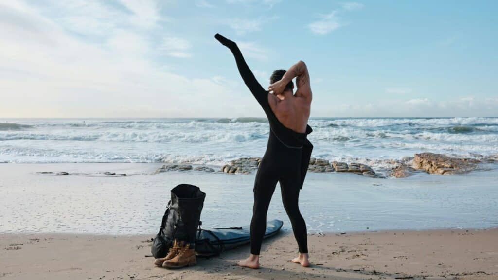 Ein Bild von einer Person, die am Strand steht und einen Neoprenanzug anzieht, mit einem Surfbrett, Stiefeln und einer Tasche (Surfzubehör) zu seiner Linken