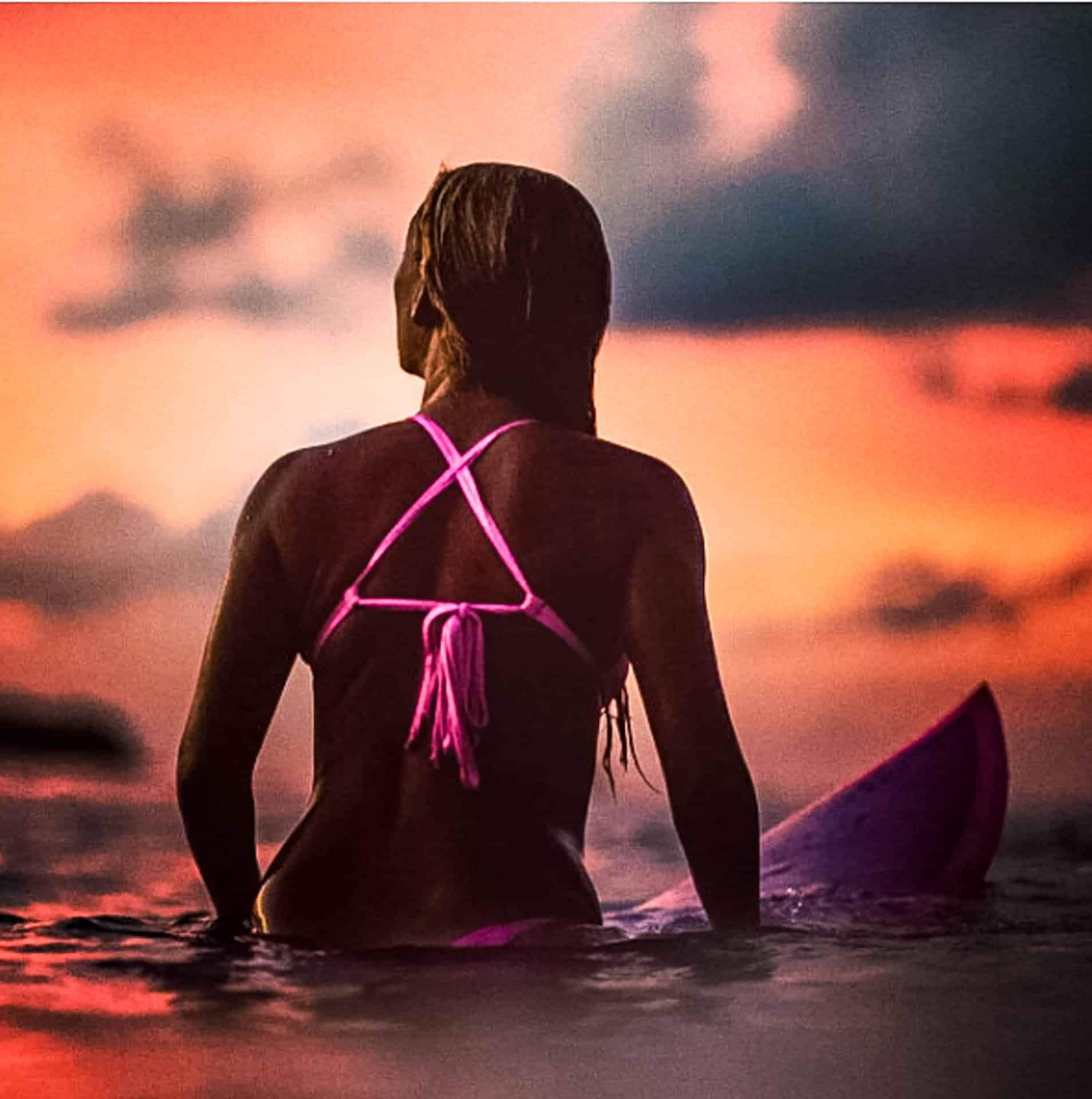 Person sitting on a surfboard looking towards the sunset.