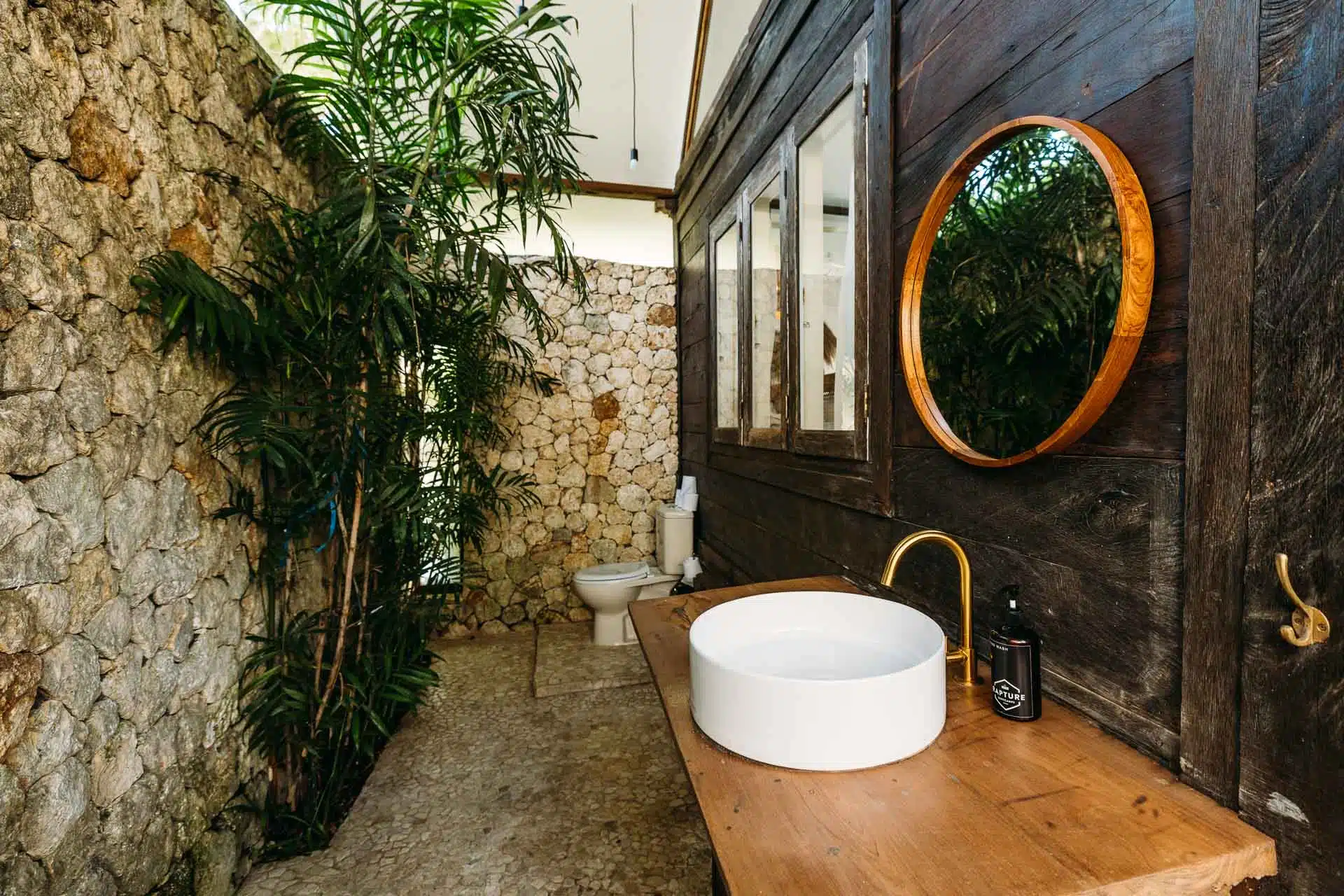 a rustic balinese outside bathroom of a surfcamp room with sink, mirror and toilet