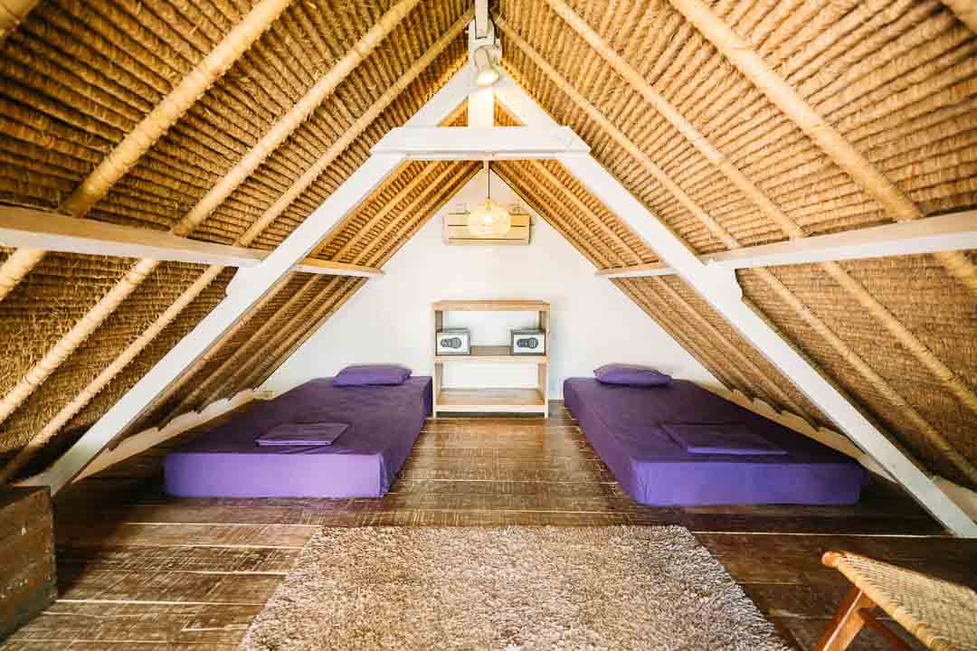 A room located at the top floor with two purple single beds in javanese roofed ceiling. Equipped with lockers and chestbox. Accented with a hanging rattan ceiling light.