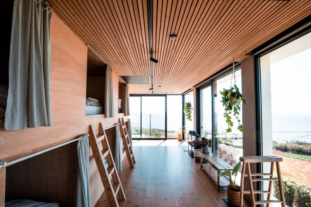 Interior of a sea-view dorm room at a surf camp, with bunk beds, wooden floors, and large windows showcasing the ocean. Surfboards and nautical decor create a cozy, beachy vibe.