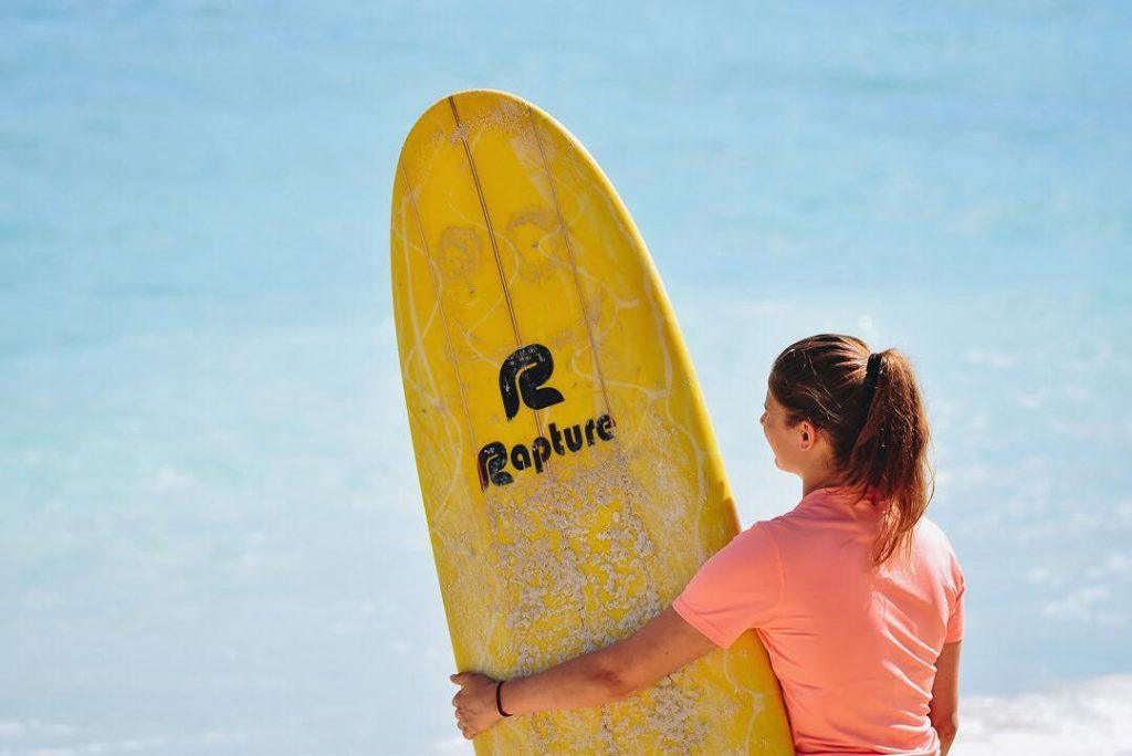 A girl holding a yellow surf board with Rapture written on it