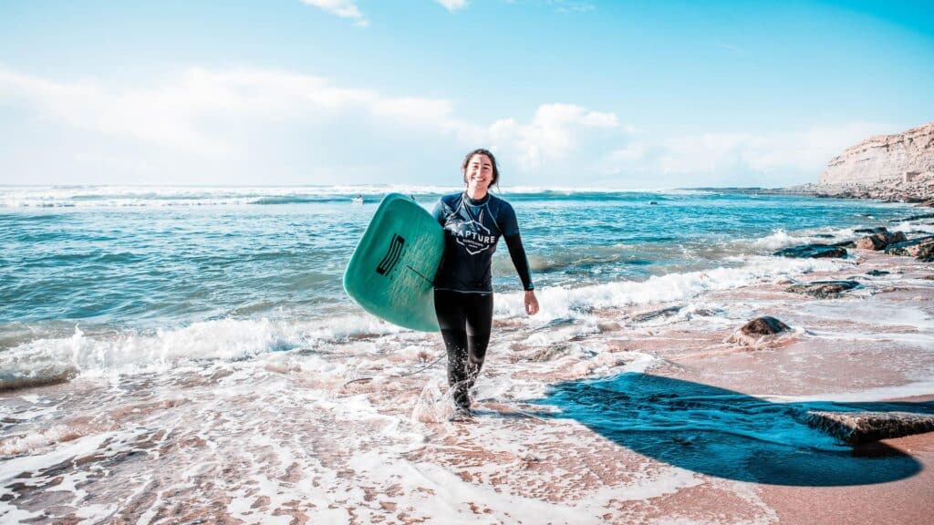 An image of a surfer leaving the sea with a surfboard under her arm