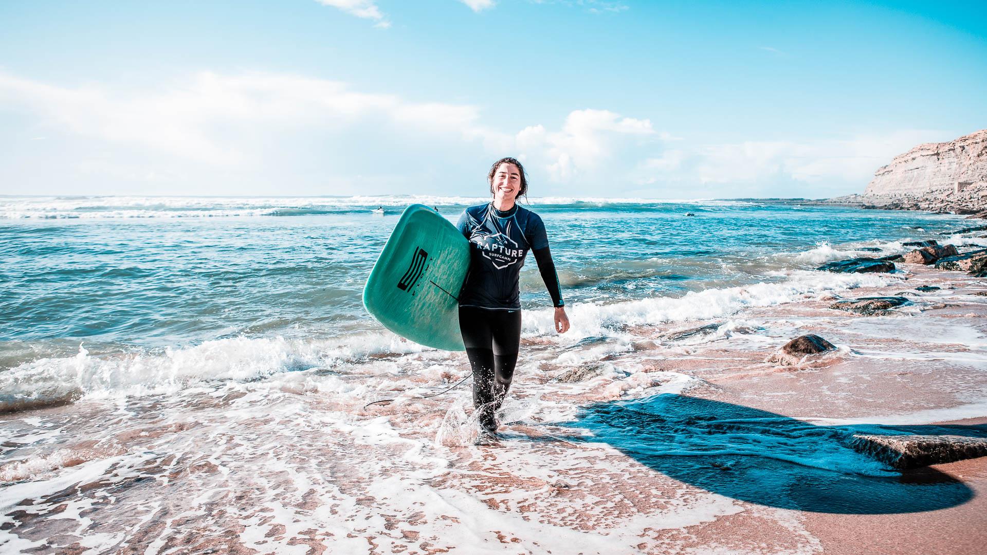 An image of a surfer leaving the sea with a surfboard under her arm