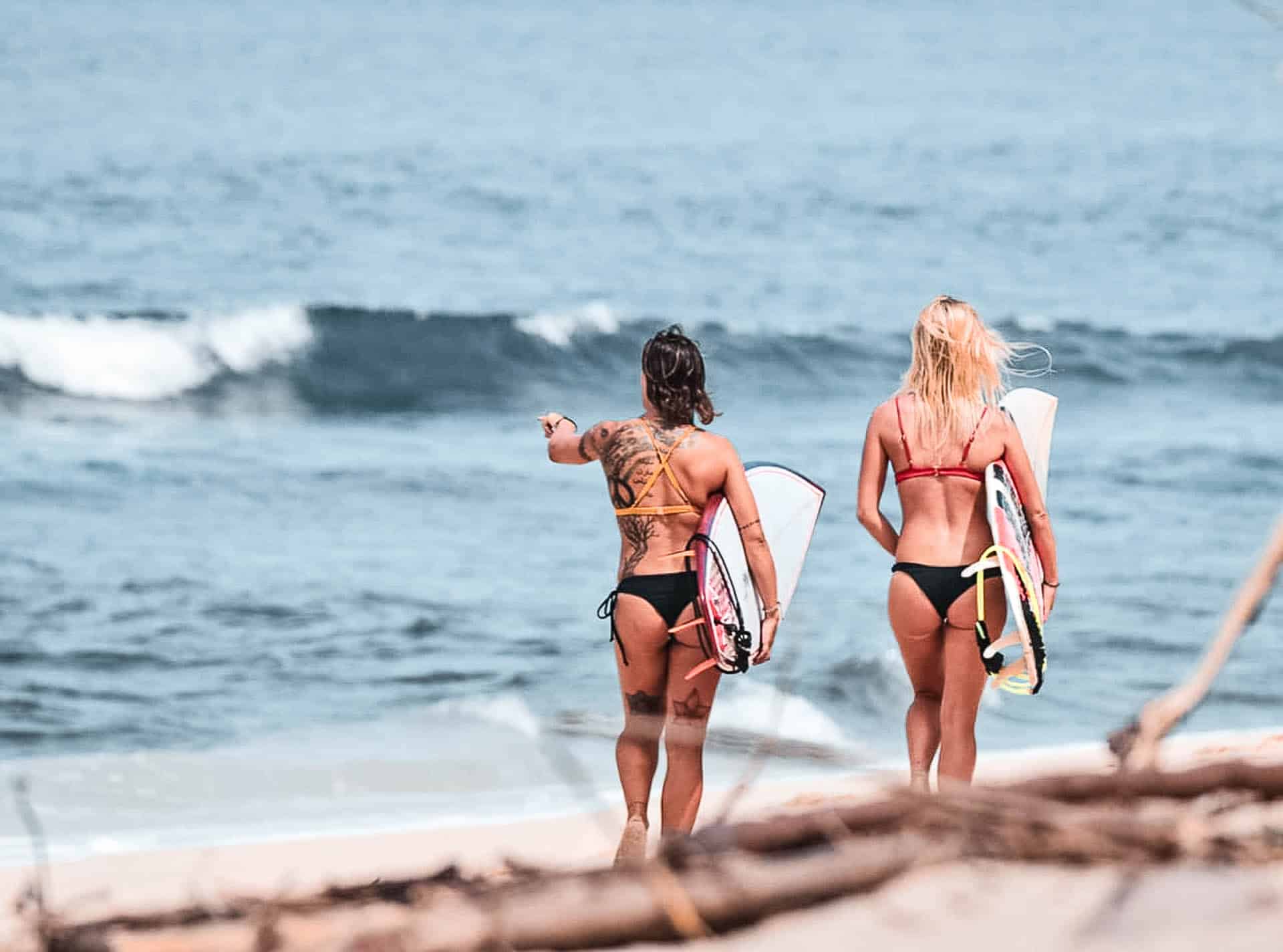 Two women in bikini walking towards the beach with surf board in hand.