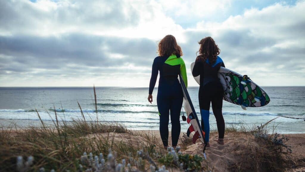 An image of two female surfers overlooking the coast of Portugal. They are wearing wetsuits and holding surfboards